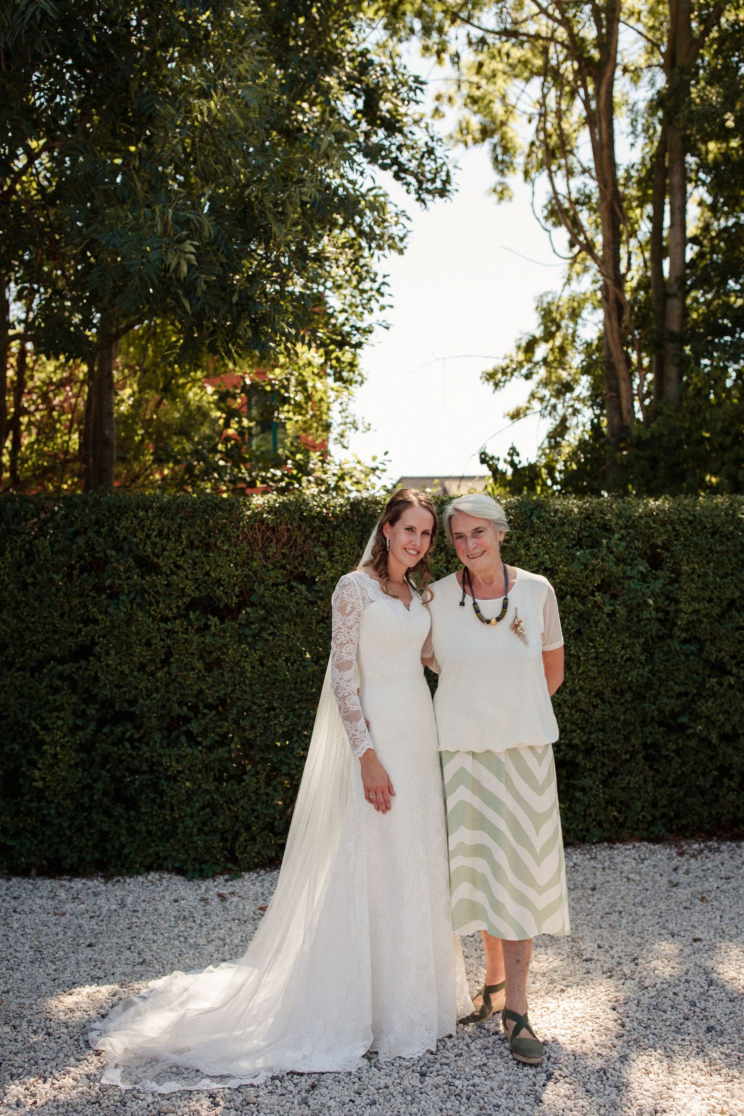 Two people in white outfits stand together on a gravel path surrounded by lush greenery at golden hour.