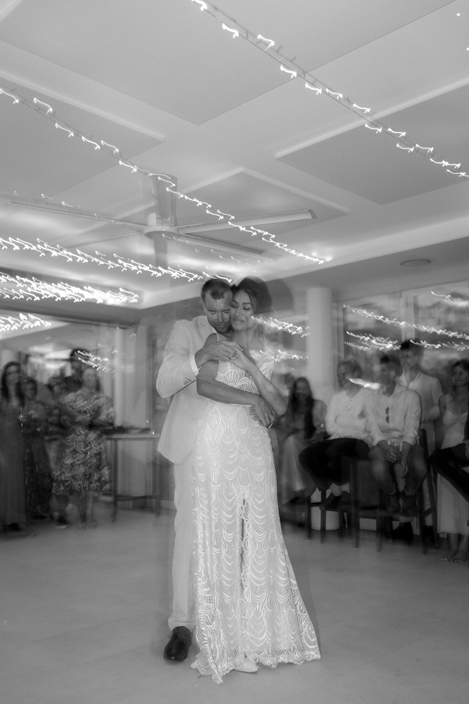 A black and white photo captures a couple dancing under twinkling string lights.