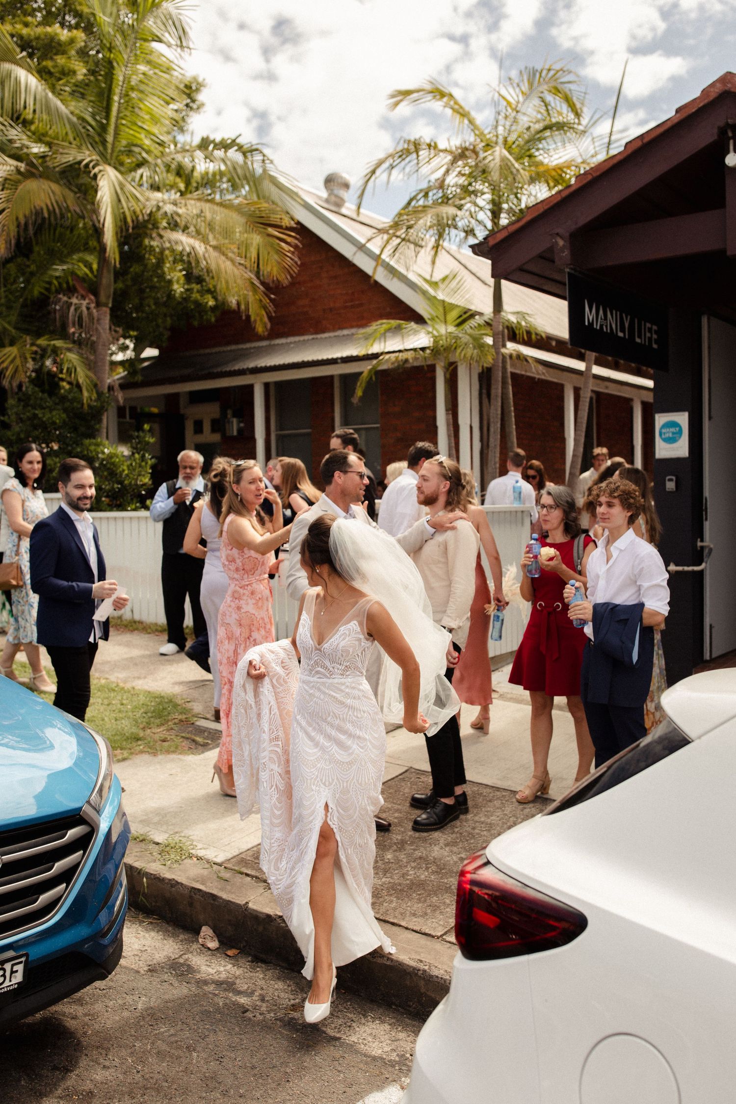 Wedding party celebrates outside brick building with palm trees overhead.