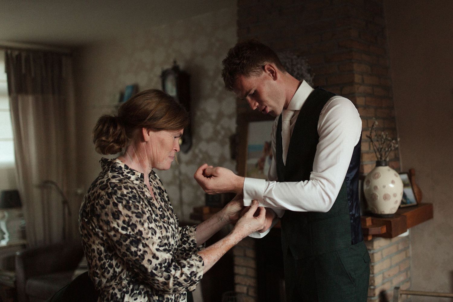 A heartfelt moment shows a mother helping with cufflinks during wedding preparations in a dimly lit room.
