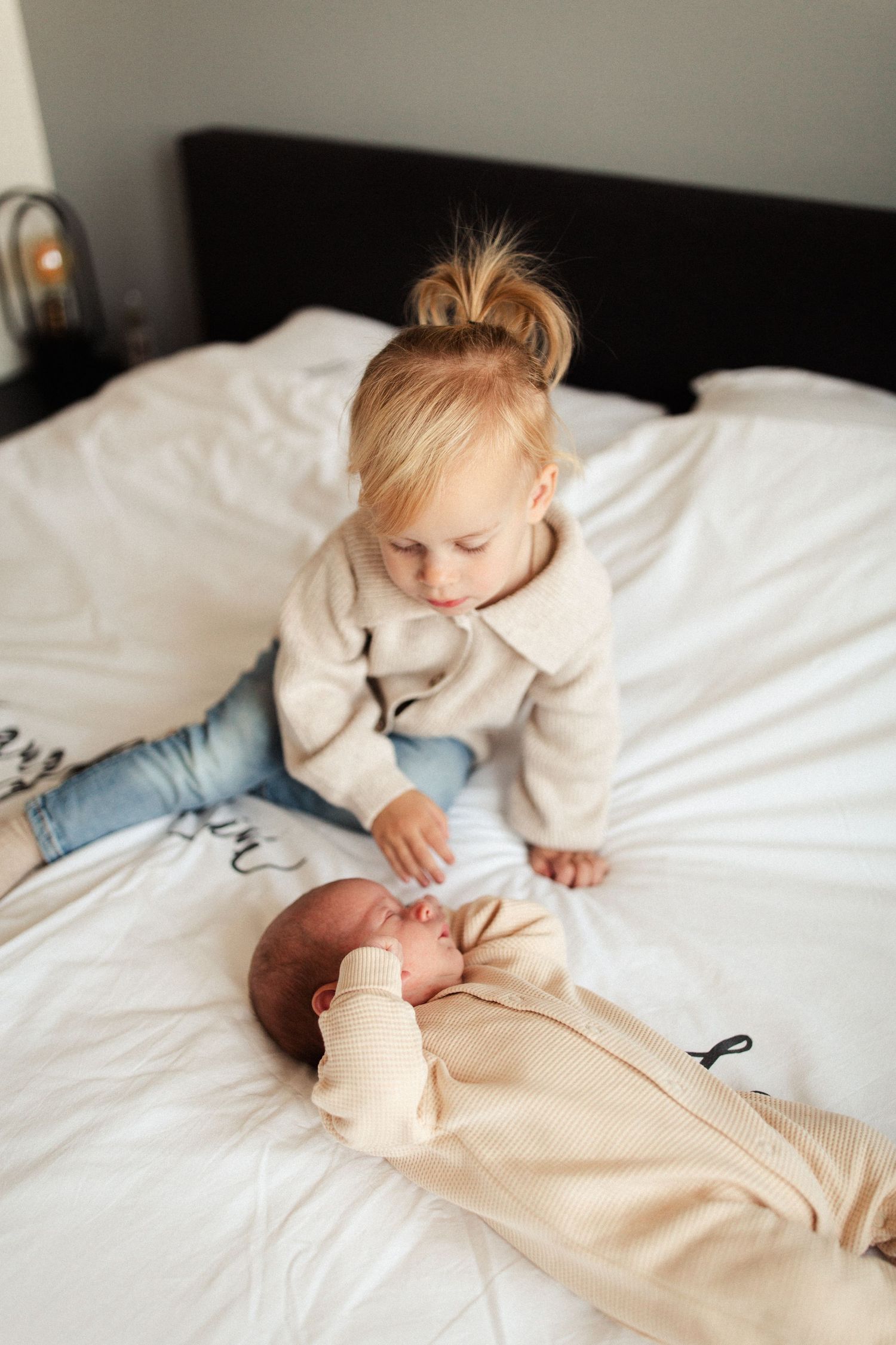 Two children in matching beige sweaters sitting together on white bedding.
