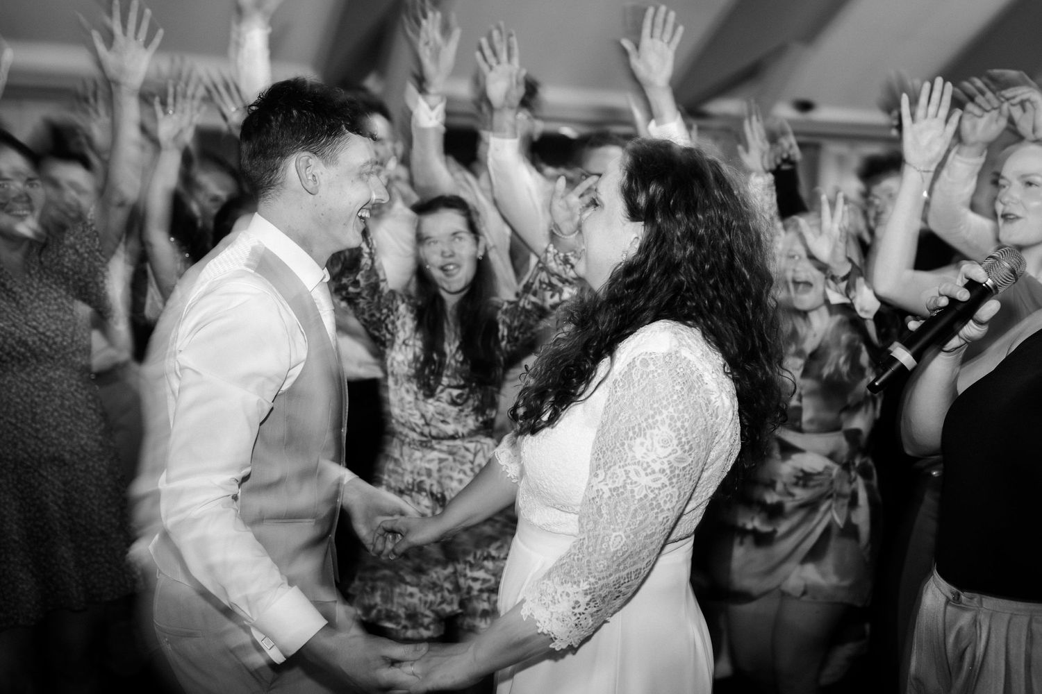 A joyful couple dances together during a wedding reception while guests raise their hands in celebration in the background.