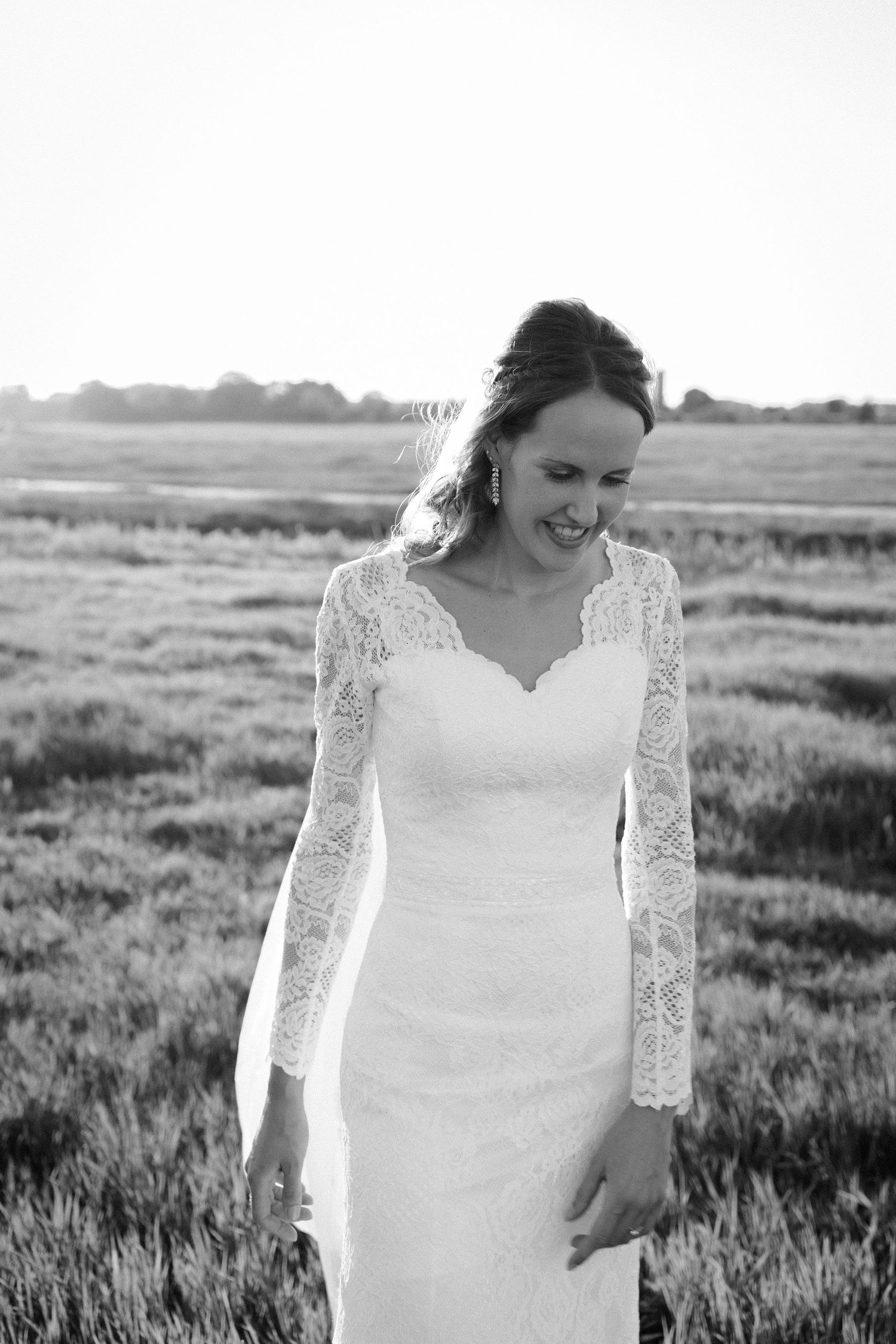 Black and white portrait of a joyful bride in a lace-sleeved wedding gown standing in a sunlit meadow.