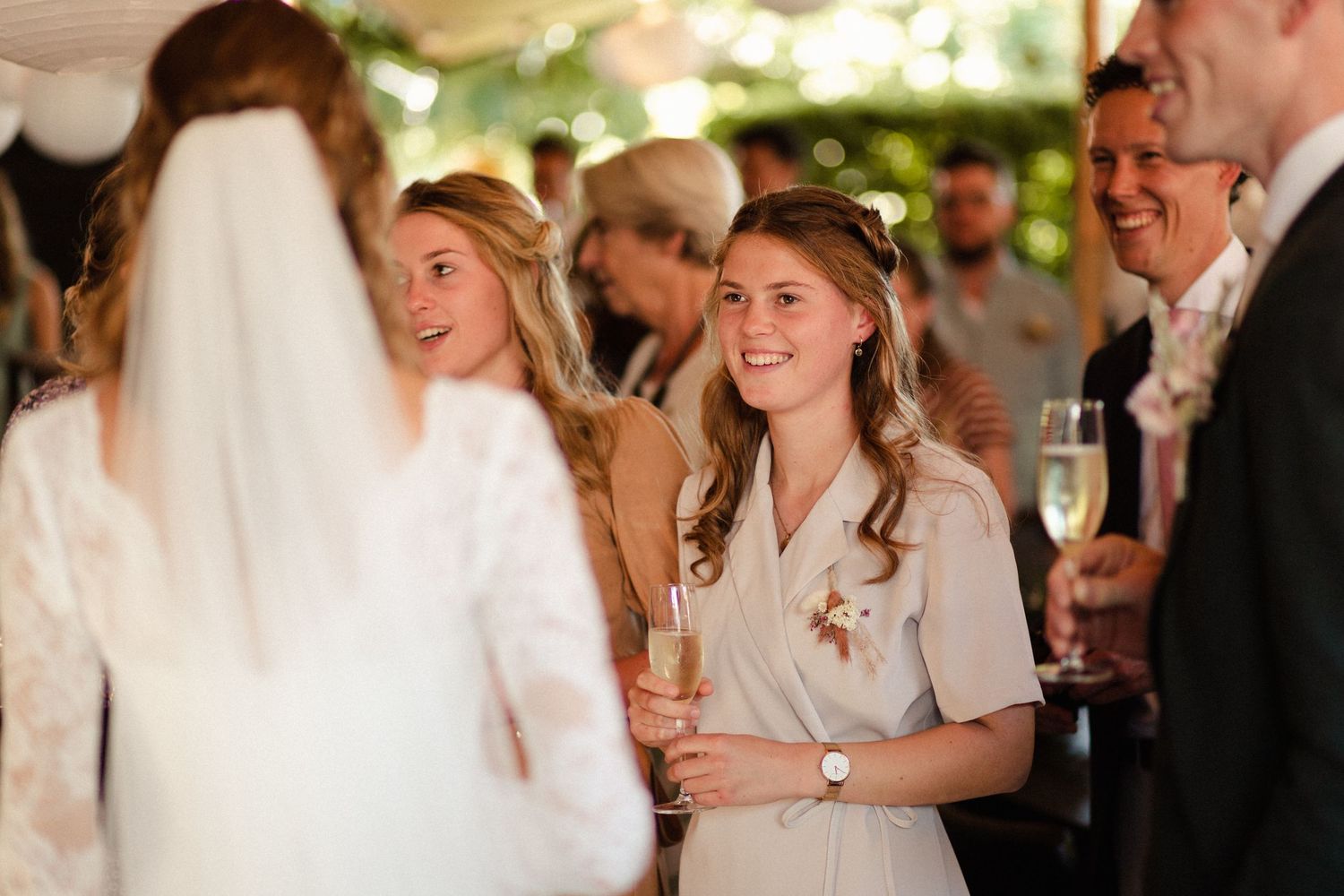 Guests in light-colored attire smile and mingle while holding champagne glasses at an elegant indoor celebration.