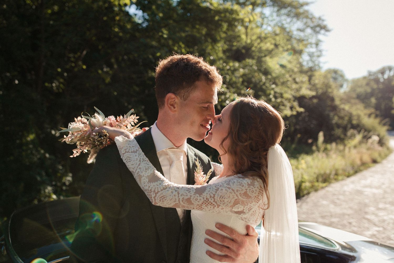 A joyful couple embraces on their wedding day with sunlit trees in the background while holding a pink bouquet.