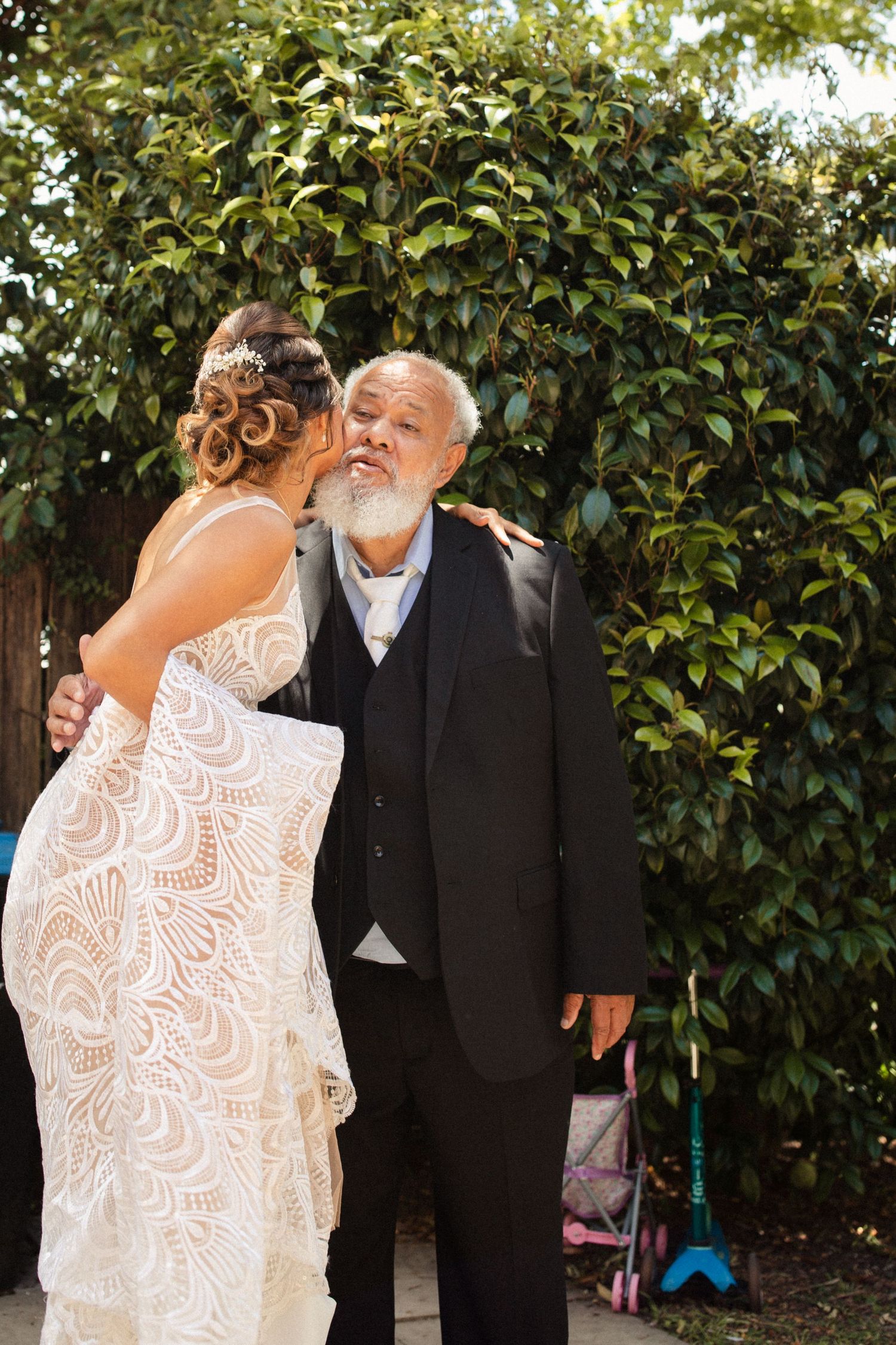 Heartwarming moment between two people in formal wedding attire near a garden hedge.