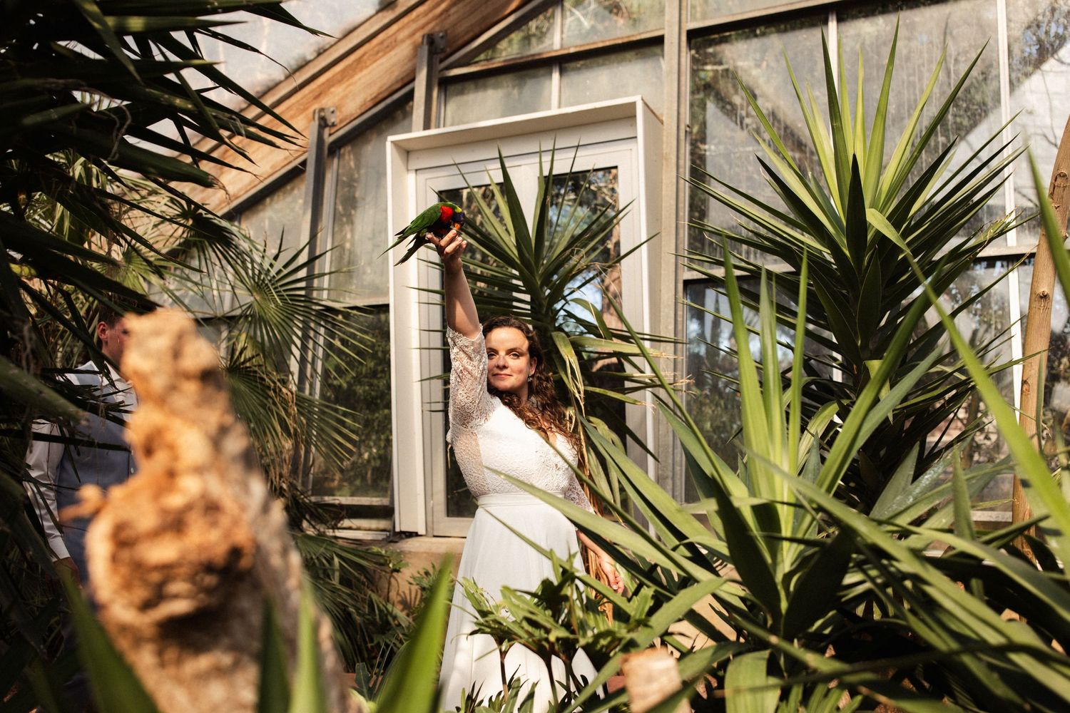A bride in a white dress poses among tropical plants and greenery outside a white building.