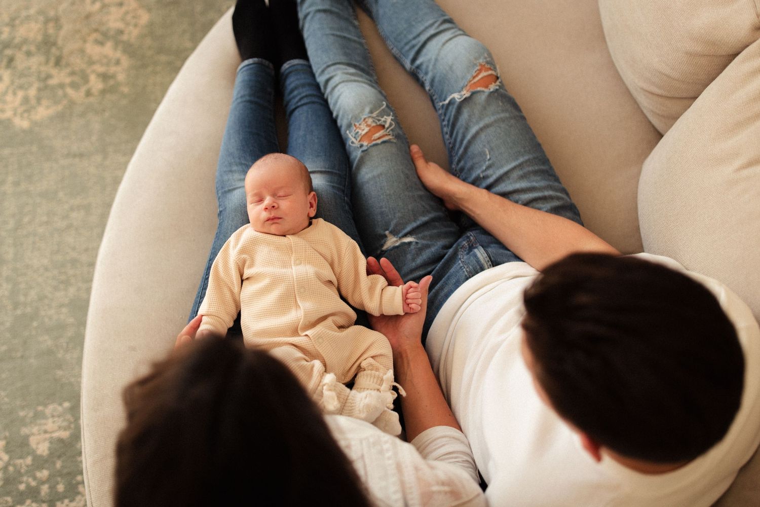 Parents relaxing on beige couch with newborn baby dressed in cream colored outfit.