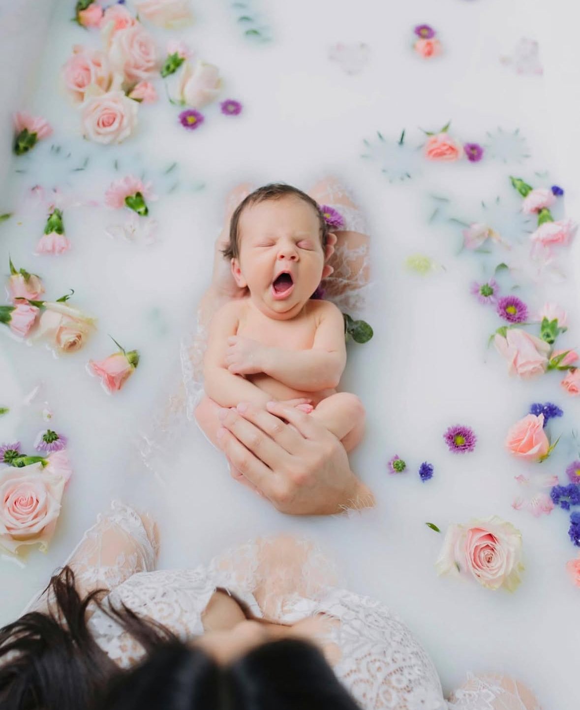 Artistic newborn photo in floral milk bath yawning while held by mom – creative newborn session by Kayleigh Kannady Photography.