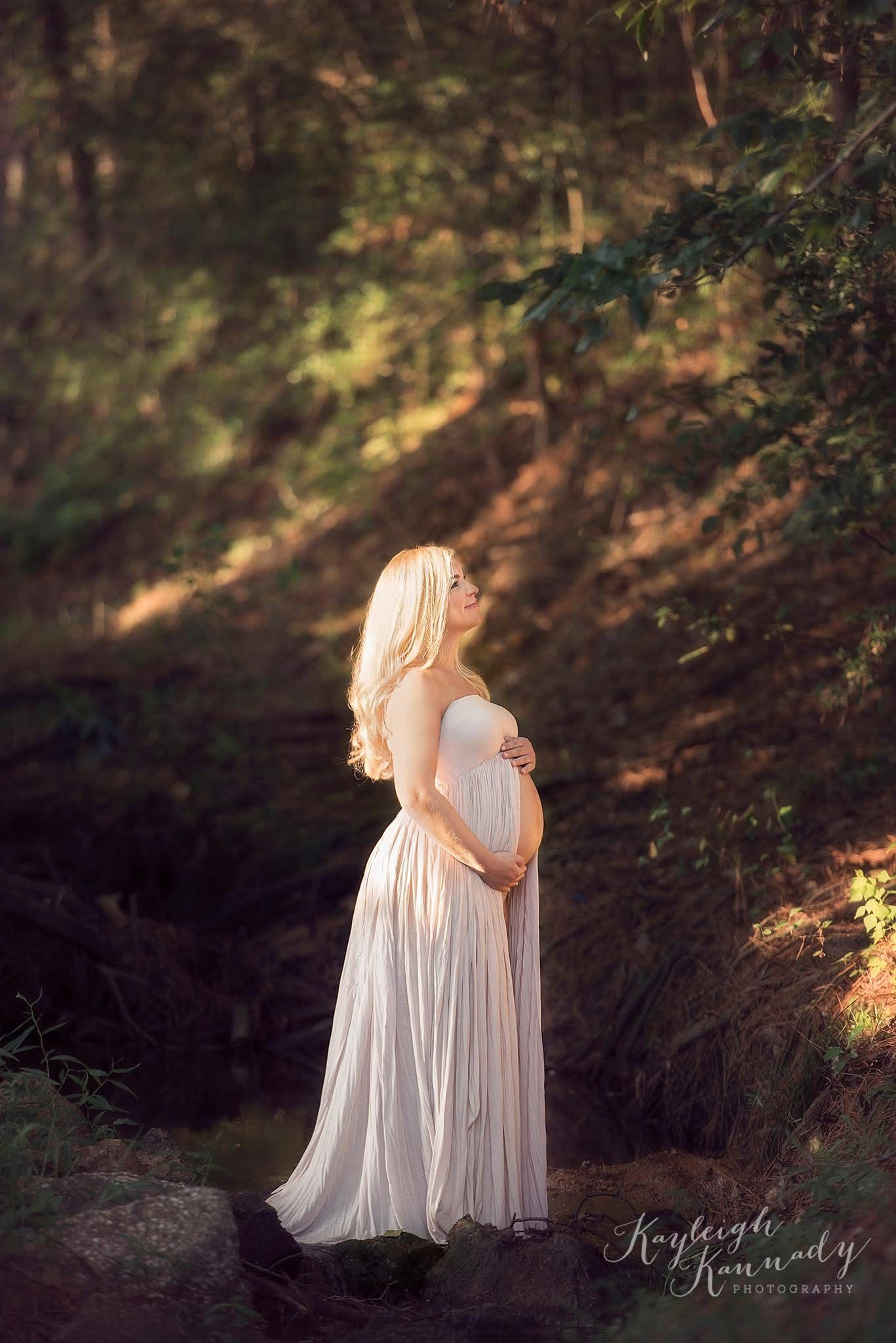 Pregnant woman in cream flowing dress at sunset in a creek holding belly – natural light maternity session by Kayleigh Kannady Photography.