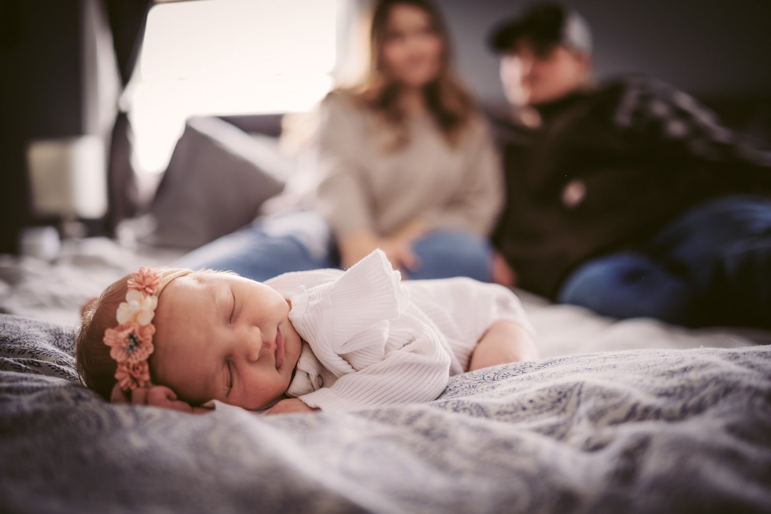 Newborn baby girl with floral headband sleeping peacefully on bed while parents sit blurred in background.