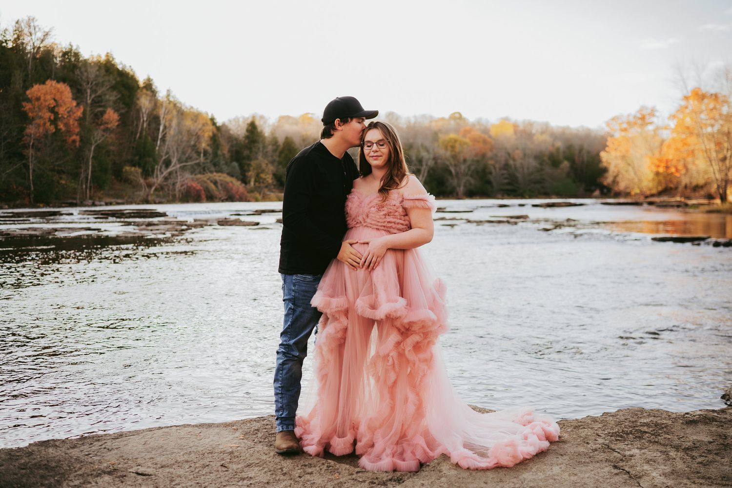 A couple in formal attire poses for romantic sunset photos along a scenic riverbank during golden hour. Maternity photographer Ontario