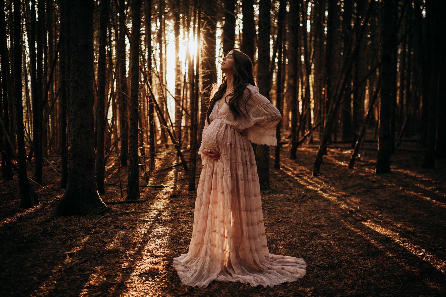 A person in a flowing pink dress stands in a sunlit forest with rays of light streaming through tall trees. Maternity photographer Ontario