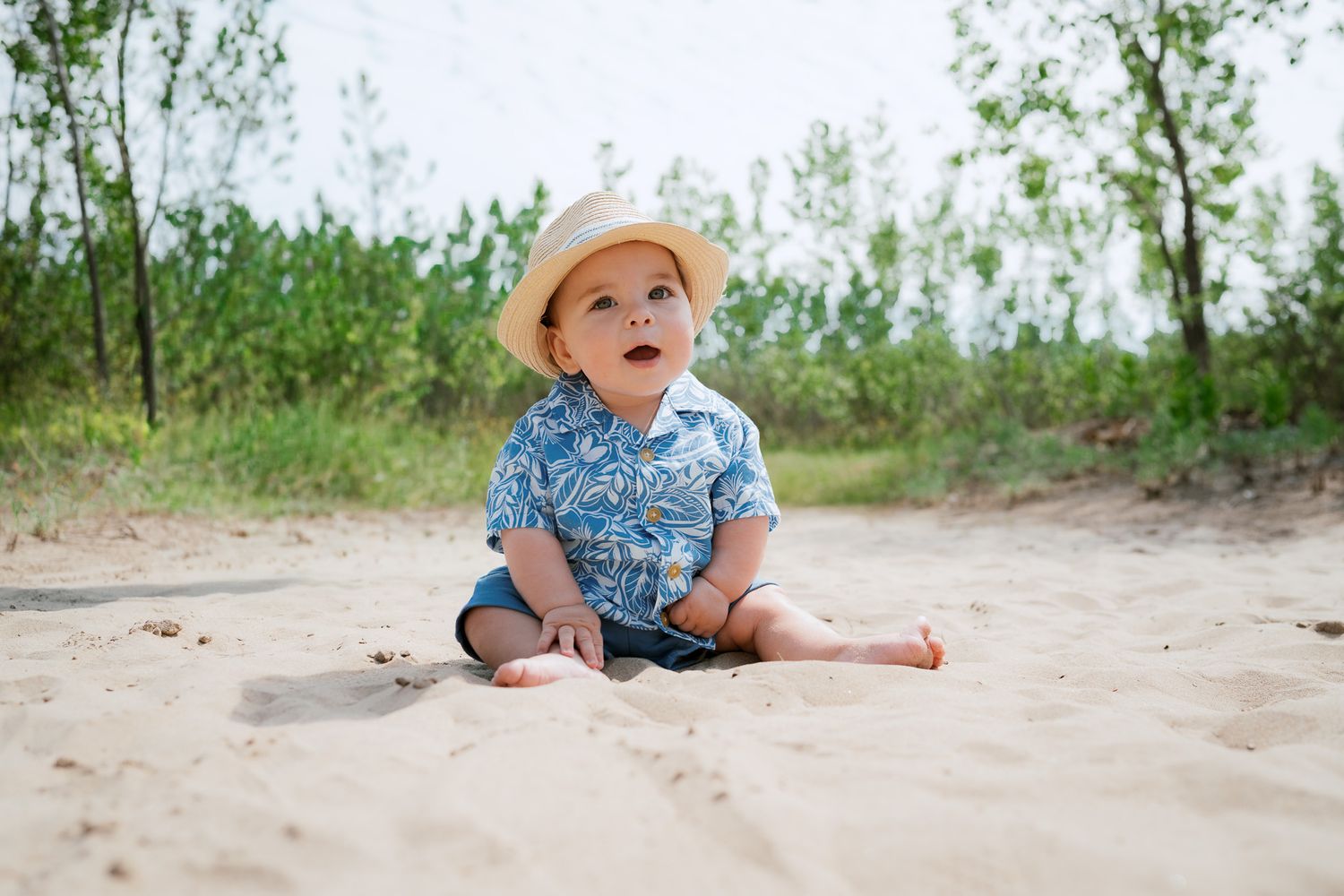 A cute baby in a straw hat and floral shirt sits on sandy beach with green trees in background.