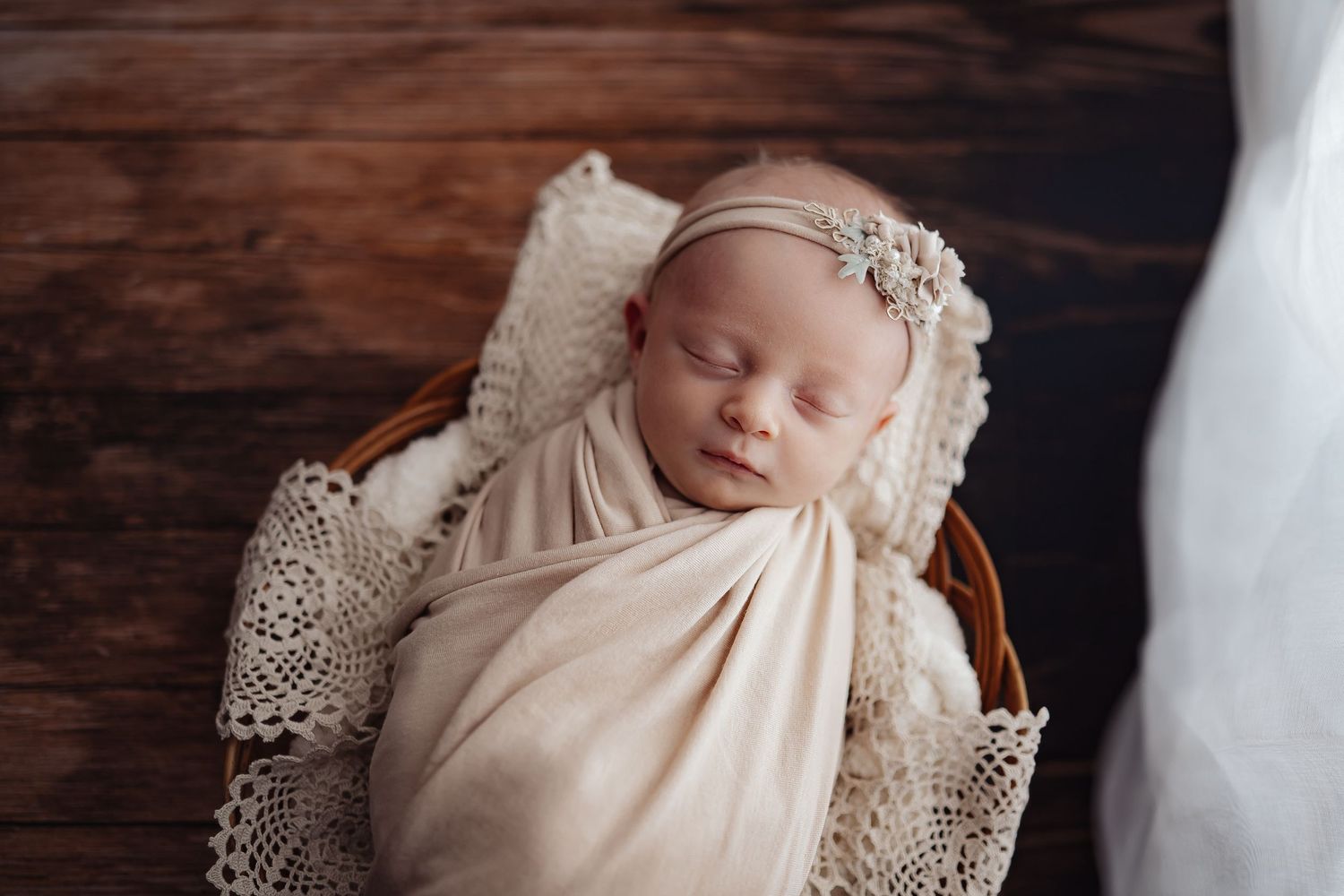 Newborn baby resting in vintage lace wrap on wooden backdrop with soft lighting.