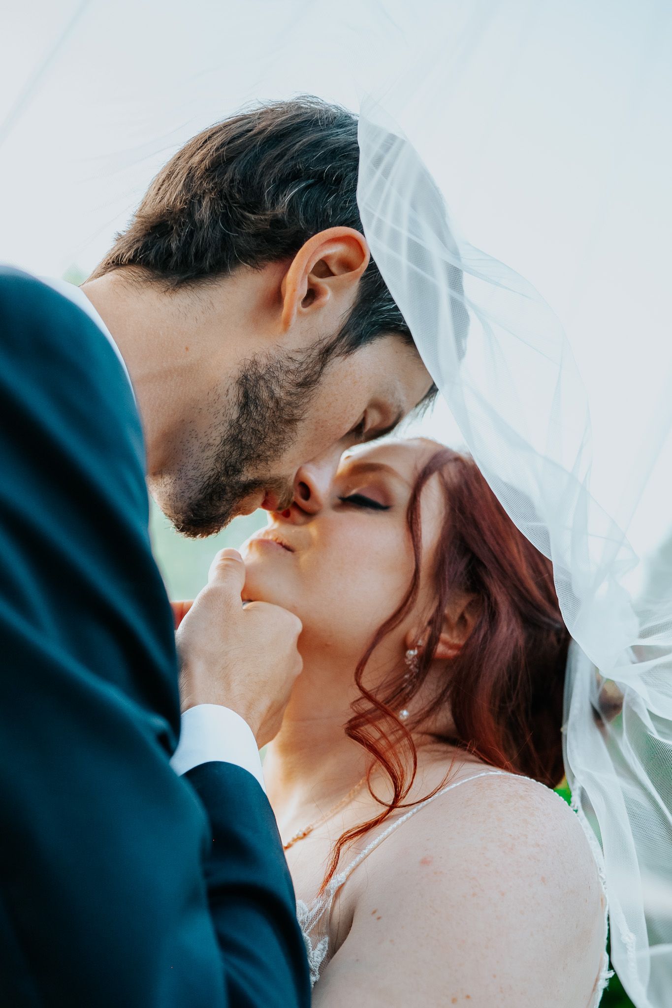 Close up of newlyweds sharing a romantic kiss under a flowing white veil during their wedding ceremony.