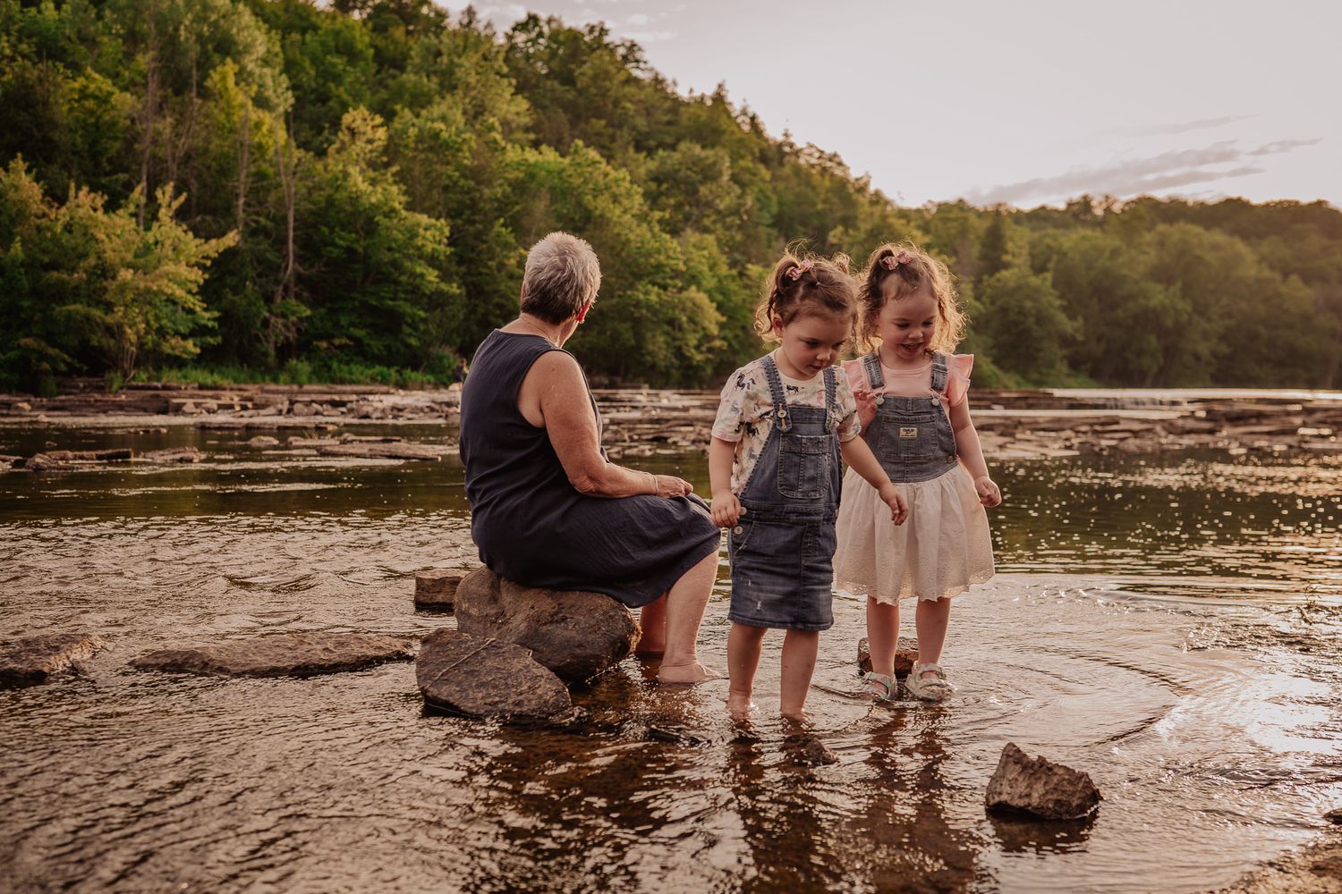 Grandmother sitting on rocks with two young girls playing in a shallow river surrounded by lush green trees at sunset.