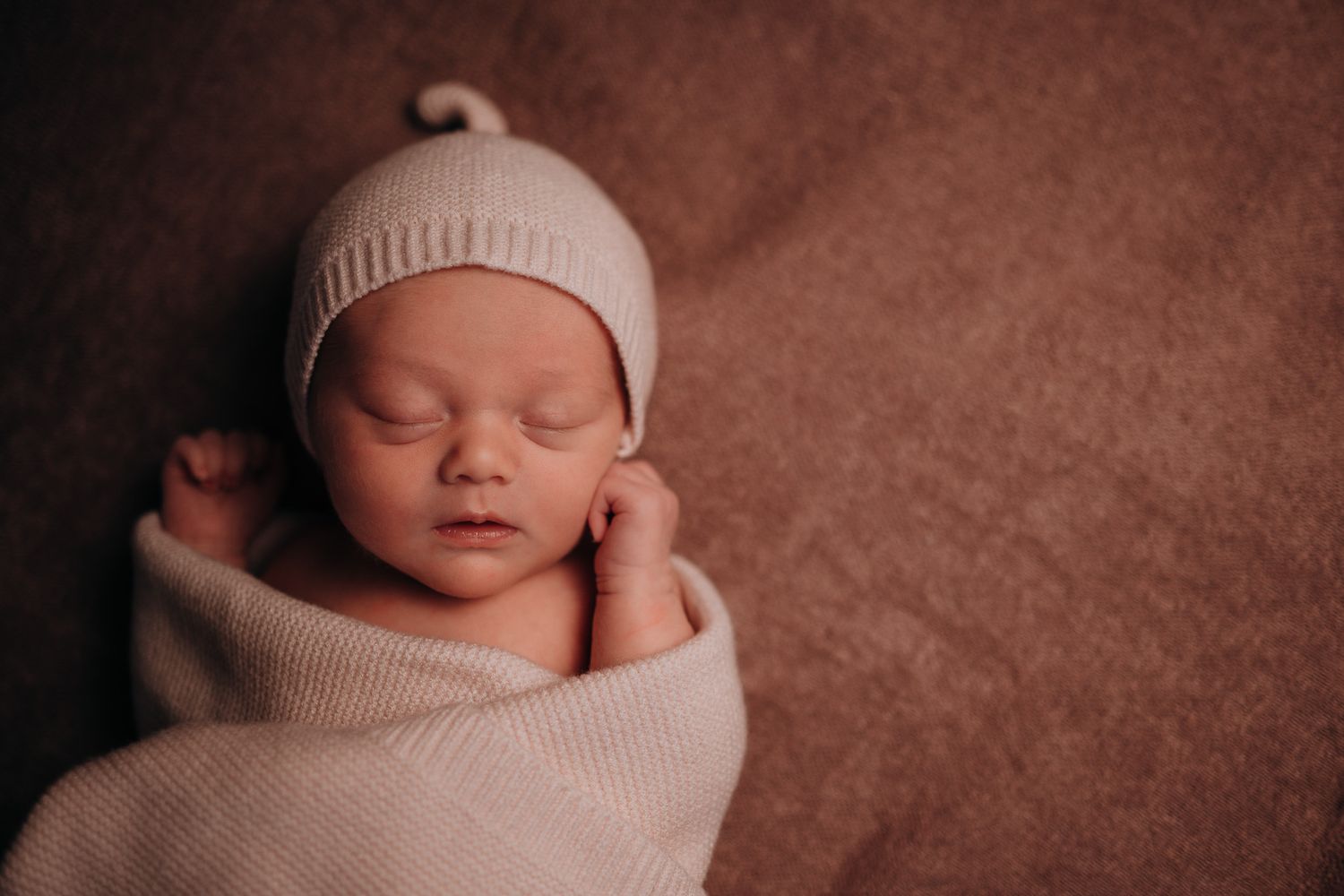 Peaceful newborn baby sleeping wrapped in a soft cream knit blanket and matching beanie hat on a warm brown background.
