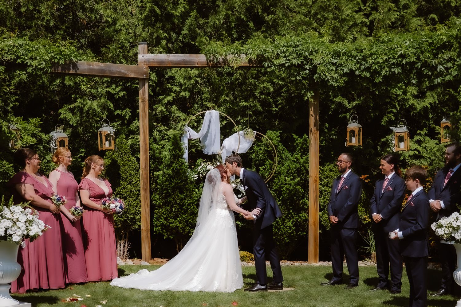 An outdoor wedding ceremony takes place in a lush garden setting with wooden posts and floral arrangements.