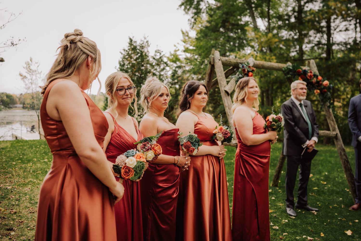 A row of bridesmaids in rust-colored dresses stand together during an outdoor wedding ceremony.