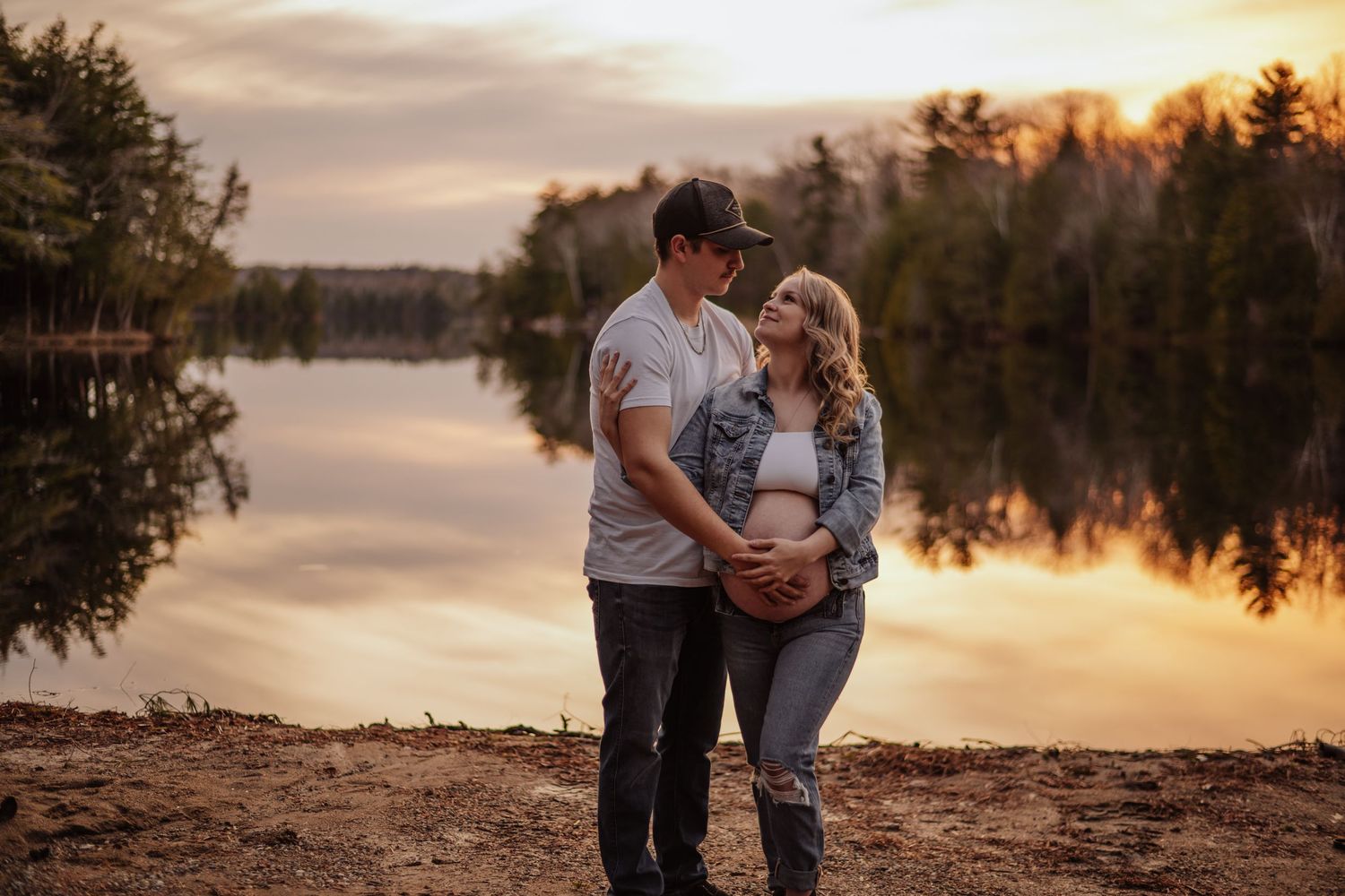 Romantic couple poses at sunset by a serene lake surrounded by trees in a warm autumn setting. Maternity photographer Ontario