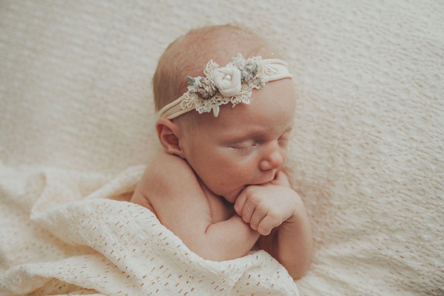 Peaceful sleeping newborn wearing lace headband on cream textured backdrop.