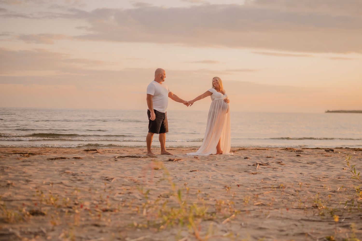 Pregnant couple holding hands on a sandy beach at golden sunset, woman in flowing white dress, man in white shirt.