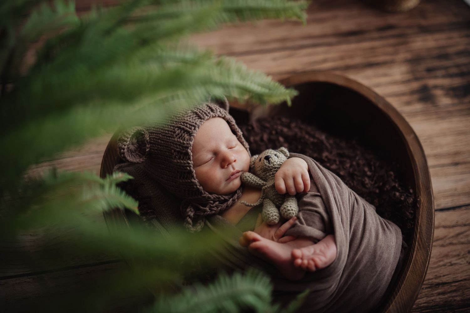 Newborn sleeping peacefully in wooden bowl surrounded by green foliage.