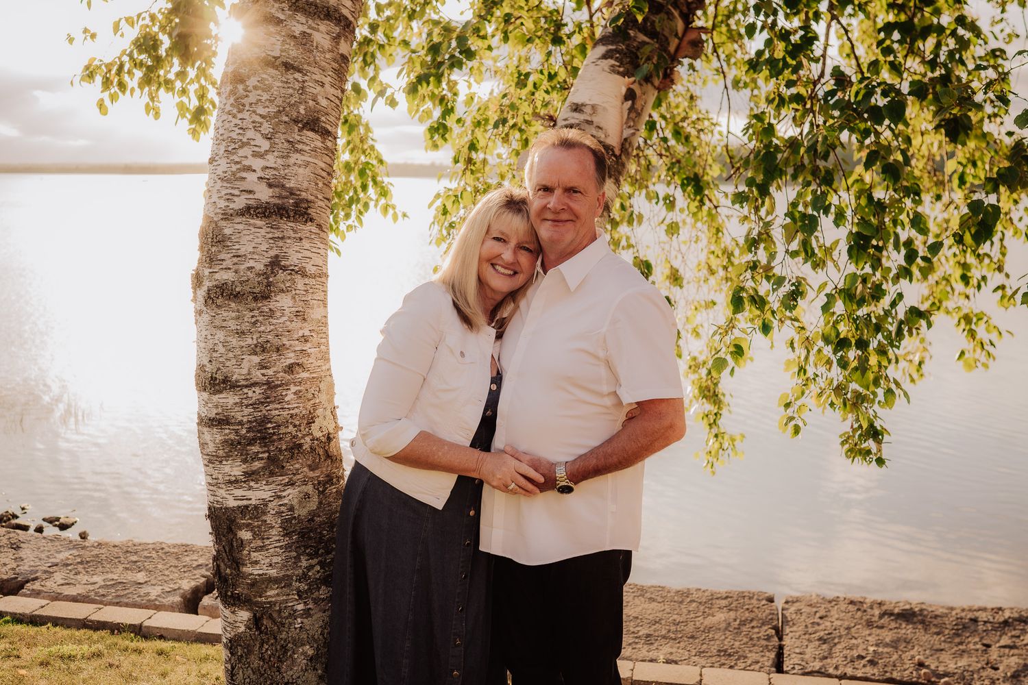 Couple embracing by a birch tree near a serene lake at sunset.