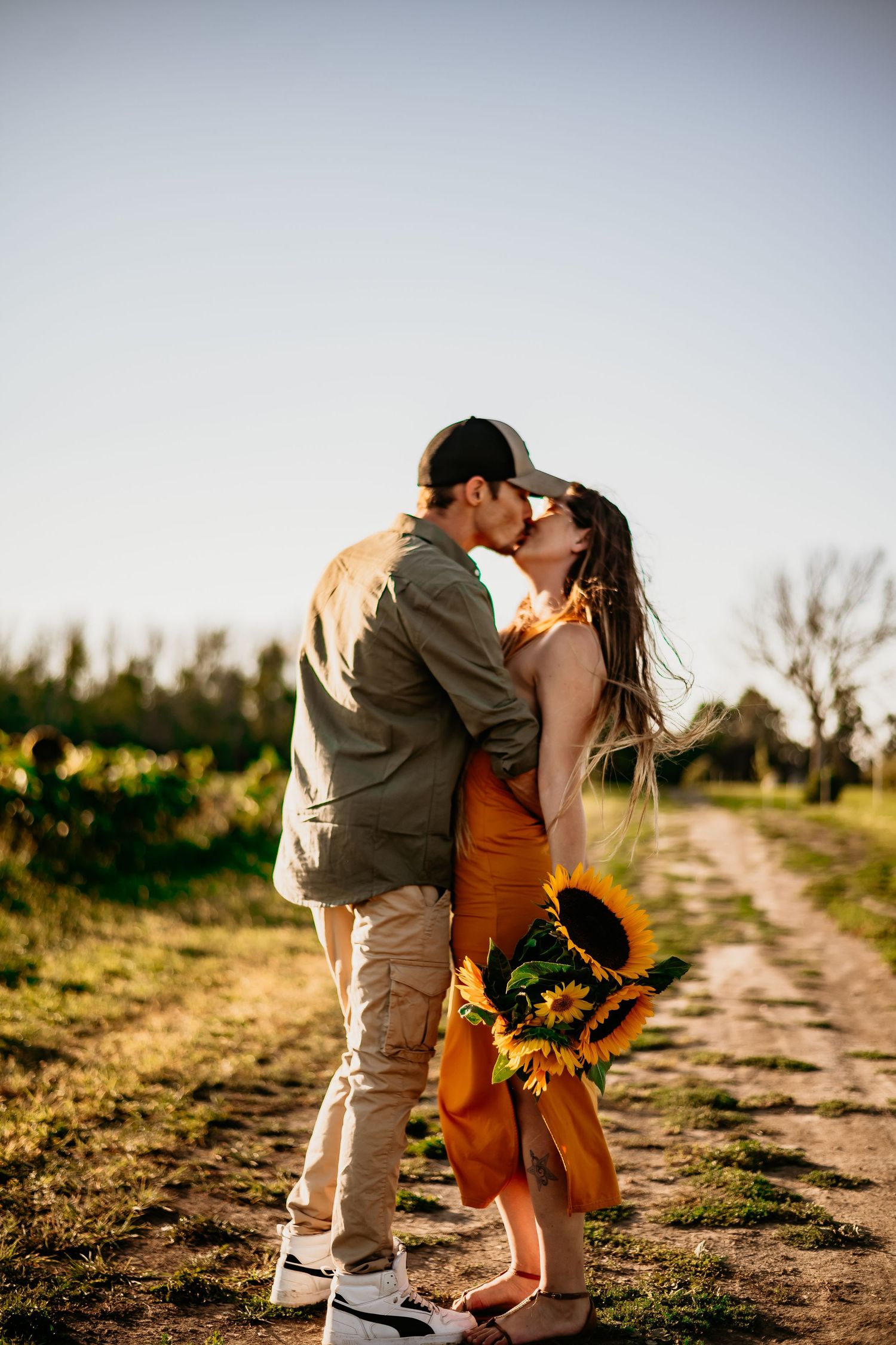 Couple kissing on a rural dirt path, woman in orange dress holding sunflowers bouquet, golden hour sunlight.
