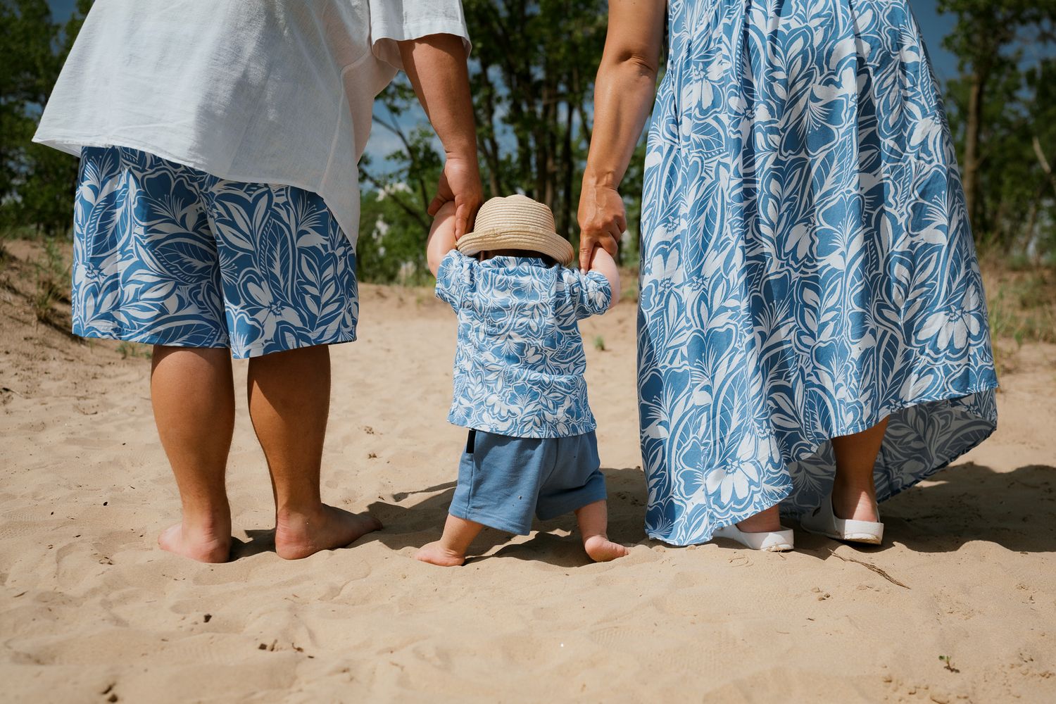 Family of three in matching blue floral outfits walking barefoot on sandy beach, holding toddler's hands.