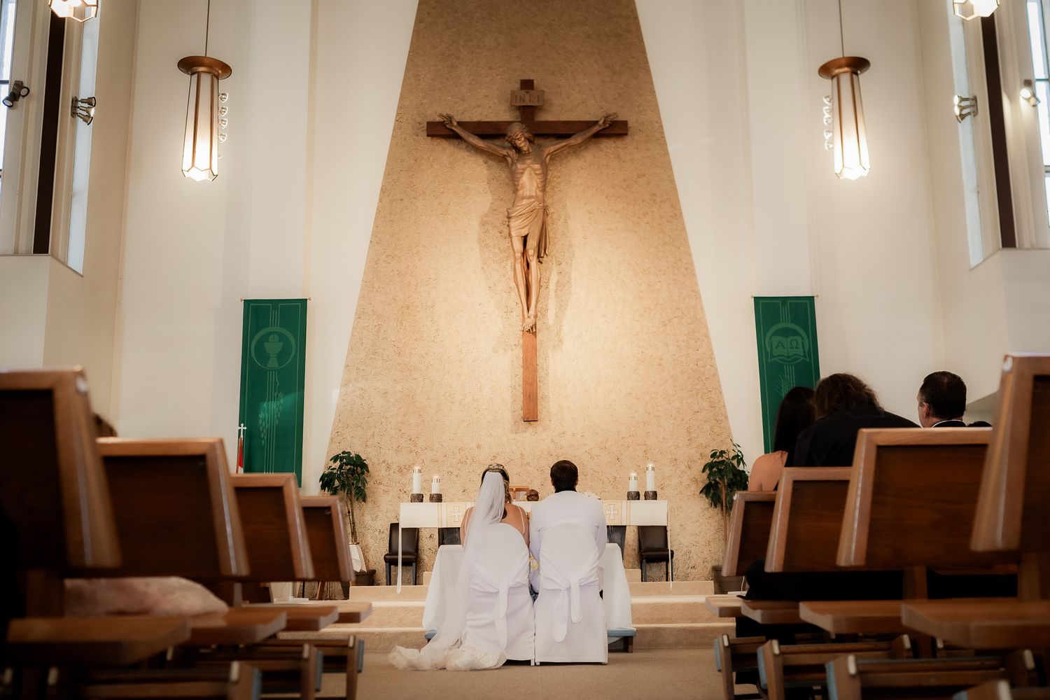 Interior of a Catholic church sanctuary with a wooden crucifix and pendant lights hanging from white vaulted ceiling.