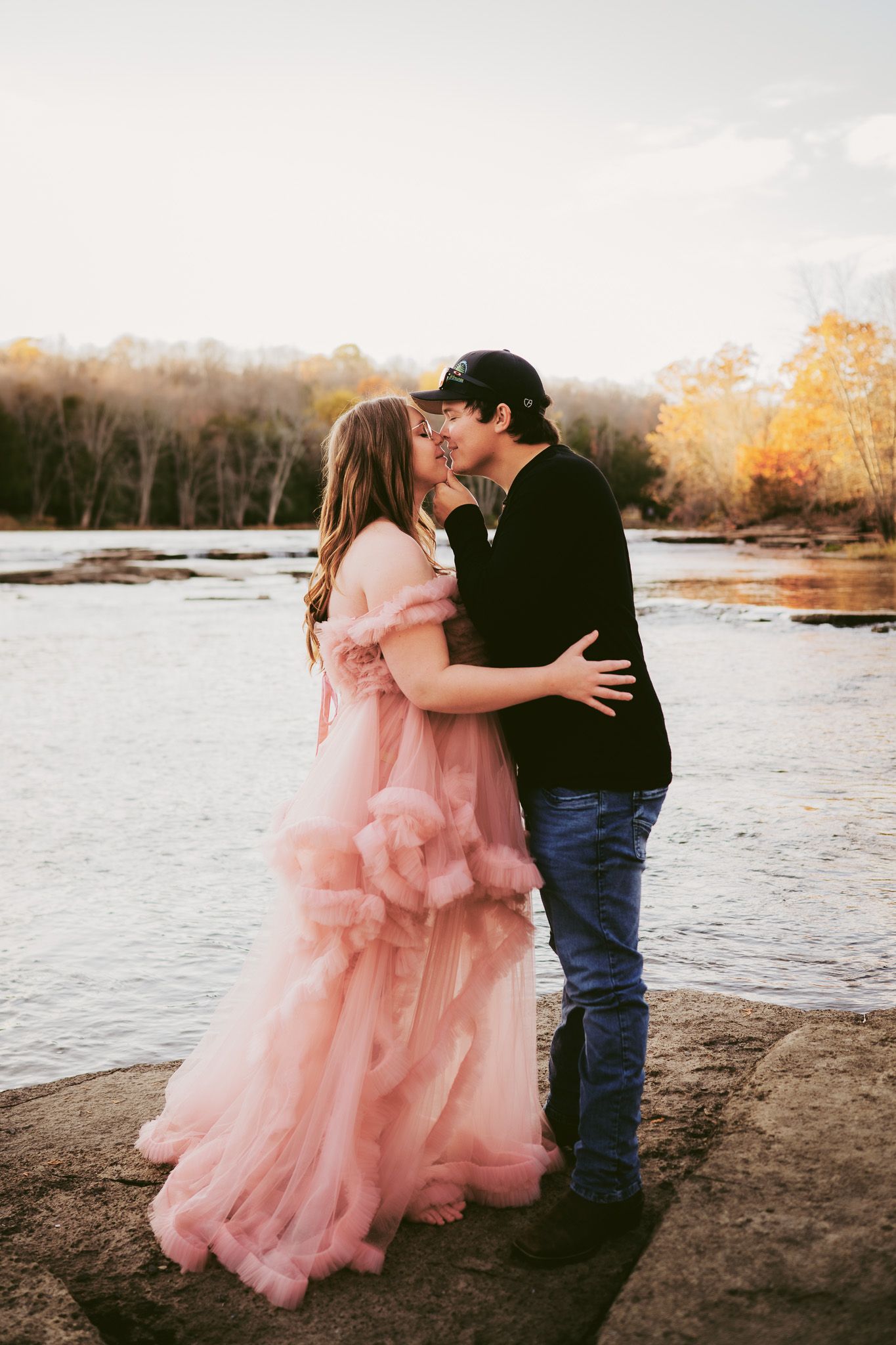 A couple shares romantic moments by a river in autumn, with the person wearing a flowing pink tulle gown. Maternity photographer Ontario