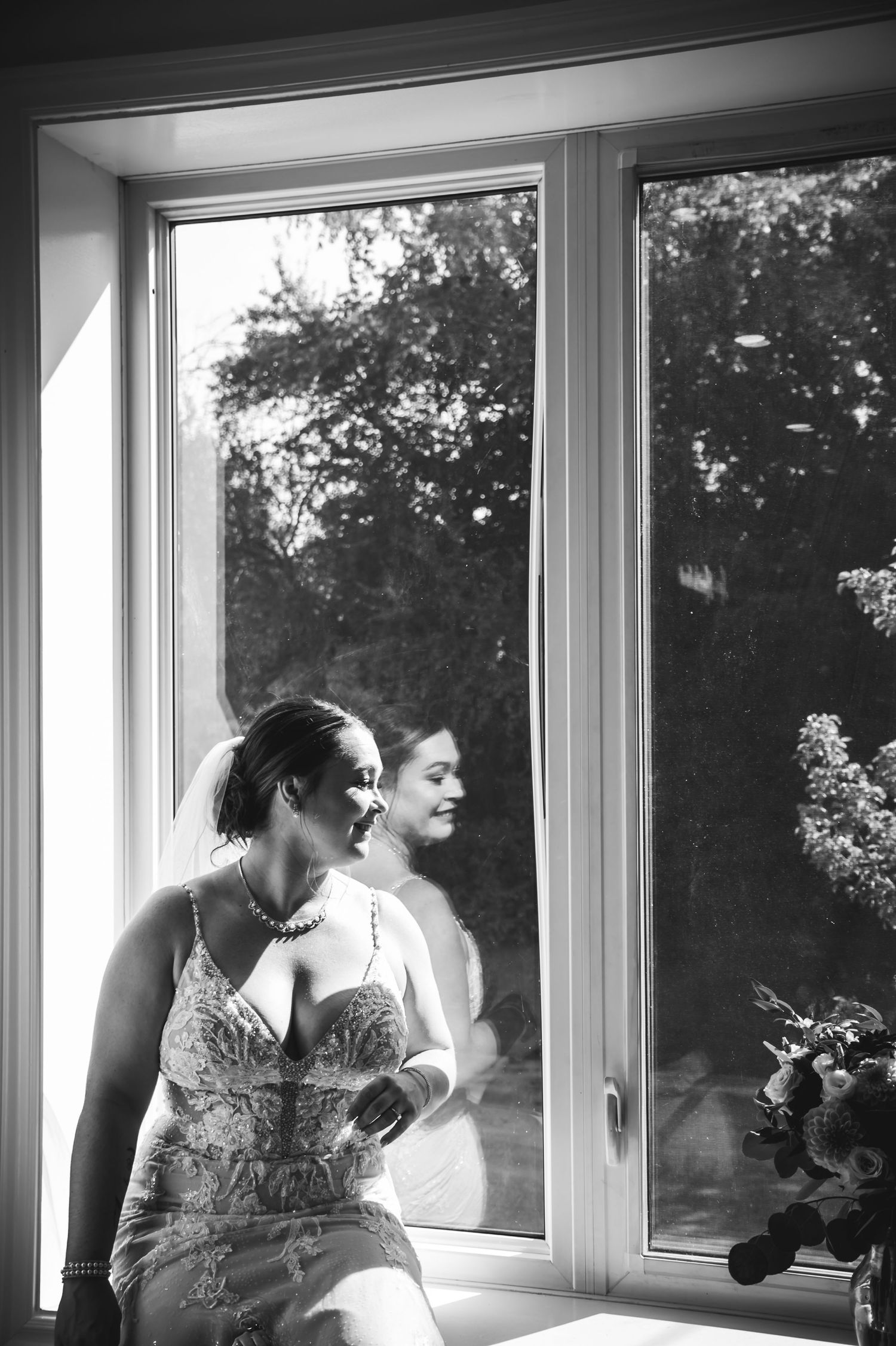 Black and white photograph of a couple in formal attire sitting by a row of windows in romantic poses.