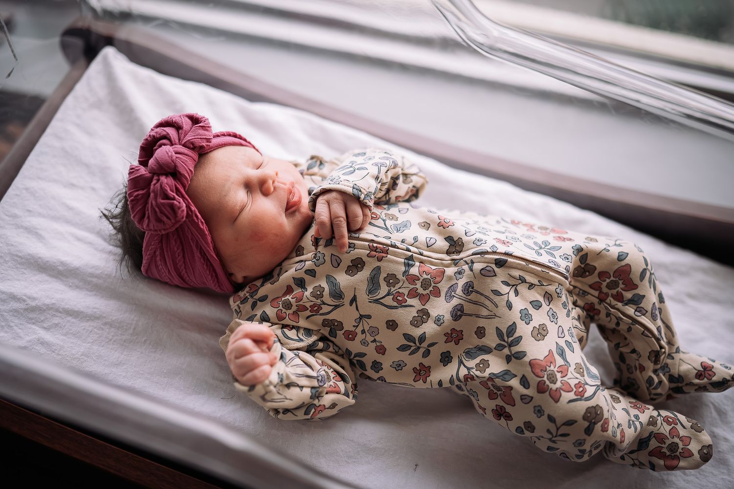 Newborn baby sleeping peacefully in a hospital bassinet wearing a floral onesie and pink bow headband.