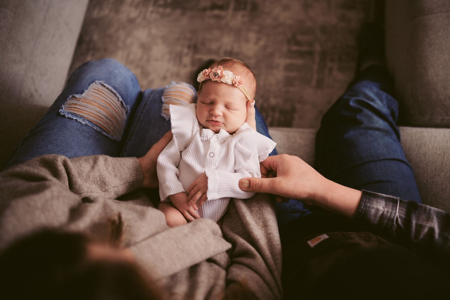 Newborn baby girl in white outfit and floral headband resting peacefully in parents arms on a cozy gray sofa.