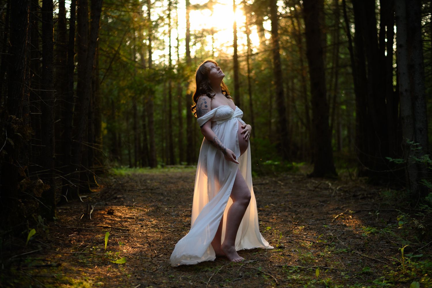 Sunlit forest path with couple in white dresses sitting together among tall evergreen trees at golden hour.