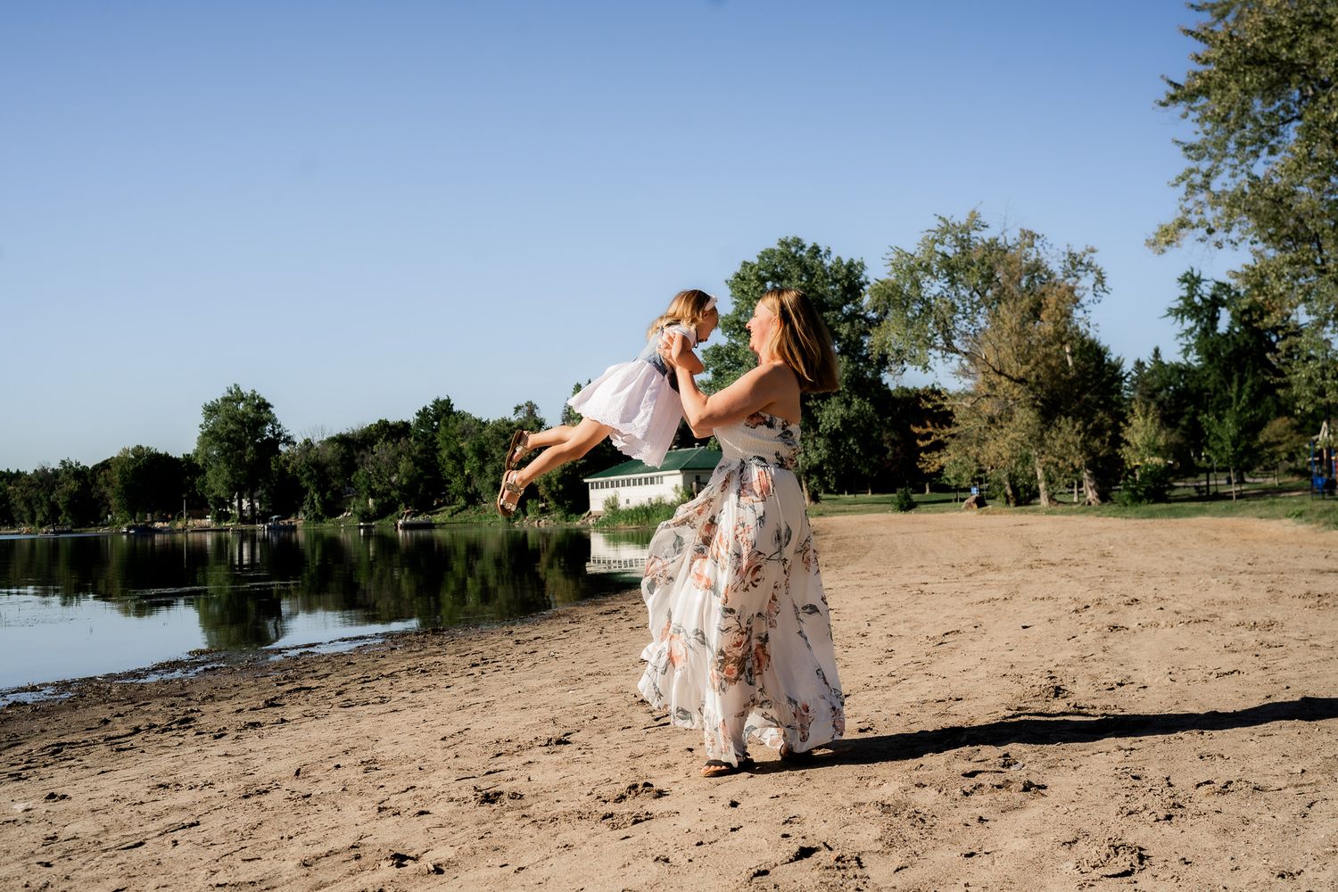 Couple in elegant attire poses for romantic photos on a sandy beach near a lake during golden hour.