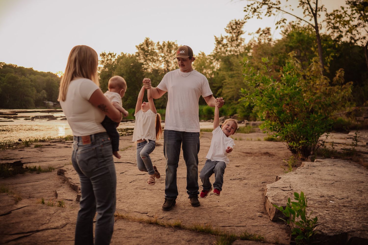 Family playing outdoors near a rocky riverbank at sunset, with dad swinging kids while mom watches joyfully.
