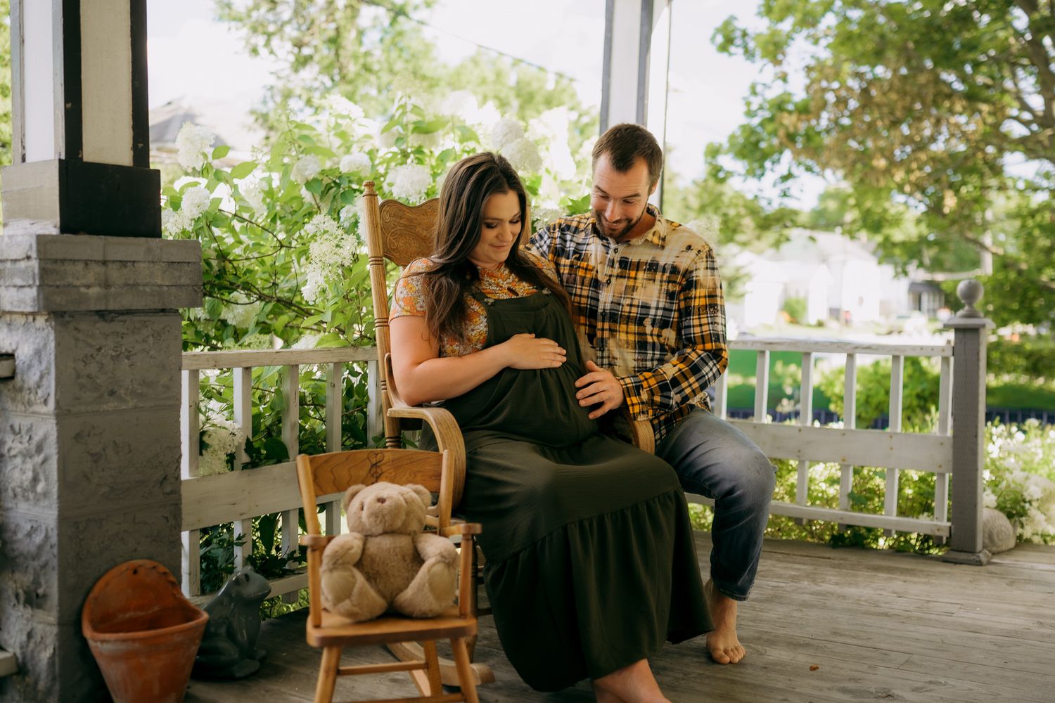 Couple sharing a tender maternity moment on a porch, man touching pregnant womans belly beside a teddy bear on a chair.