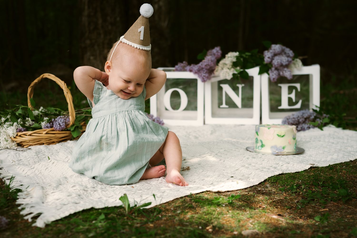 Baby girl in mint dress and party hat sits on blanket outdoors celebrating her first birthday with ONE blocks.