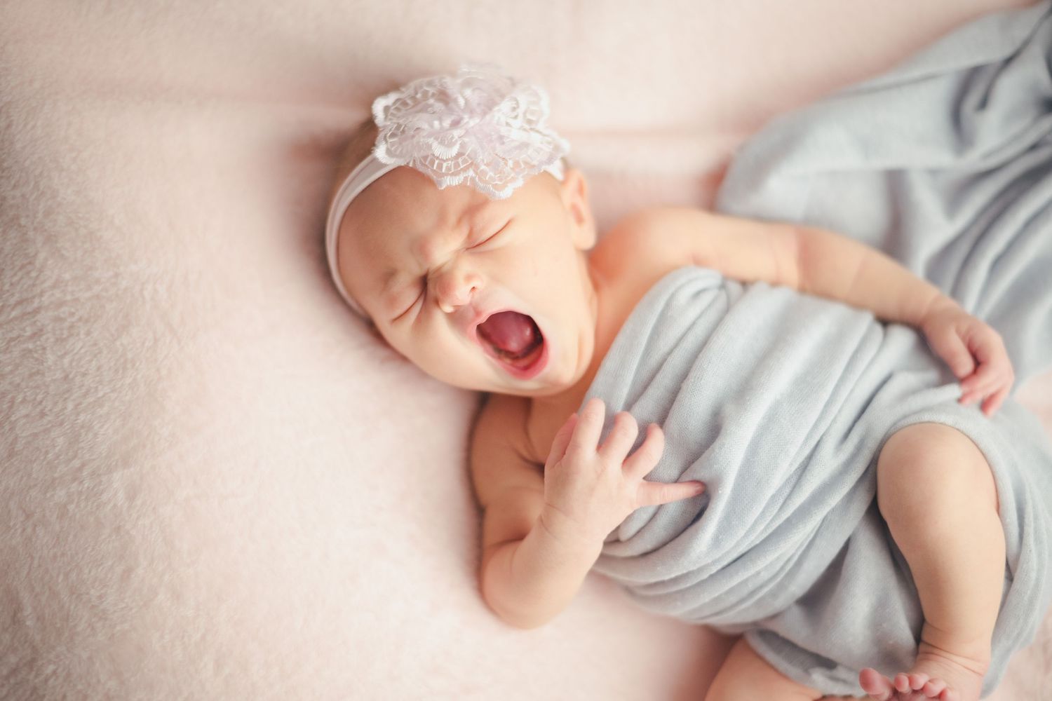 Adorable newborn baby girl yawning, wrapped in soft gray fabric, wearing a white lace headband, lying on pink blanket.