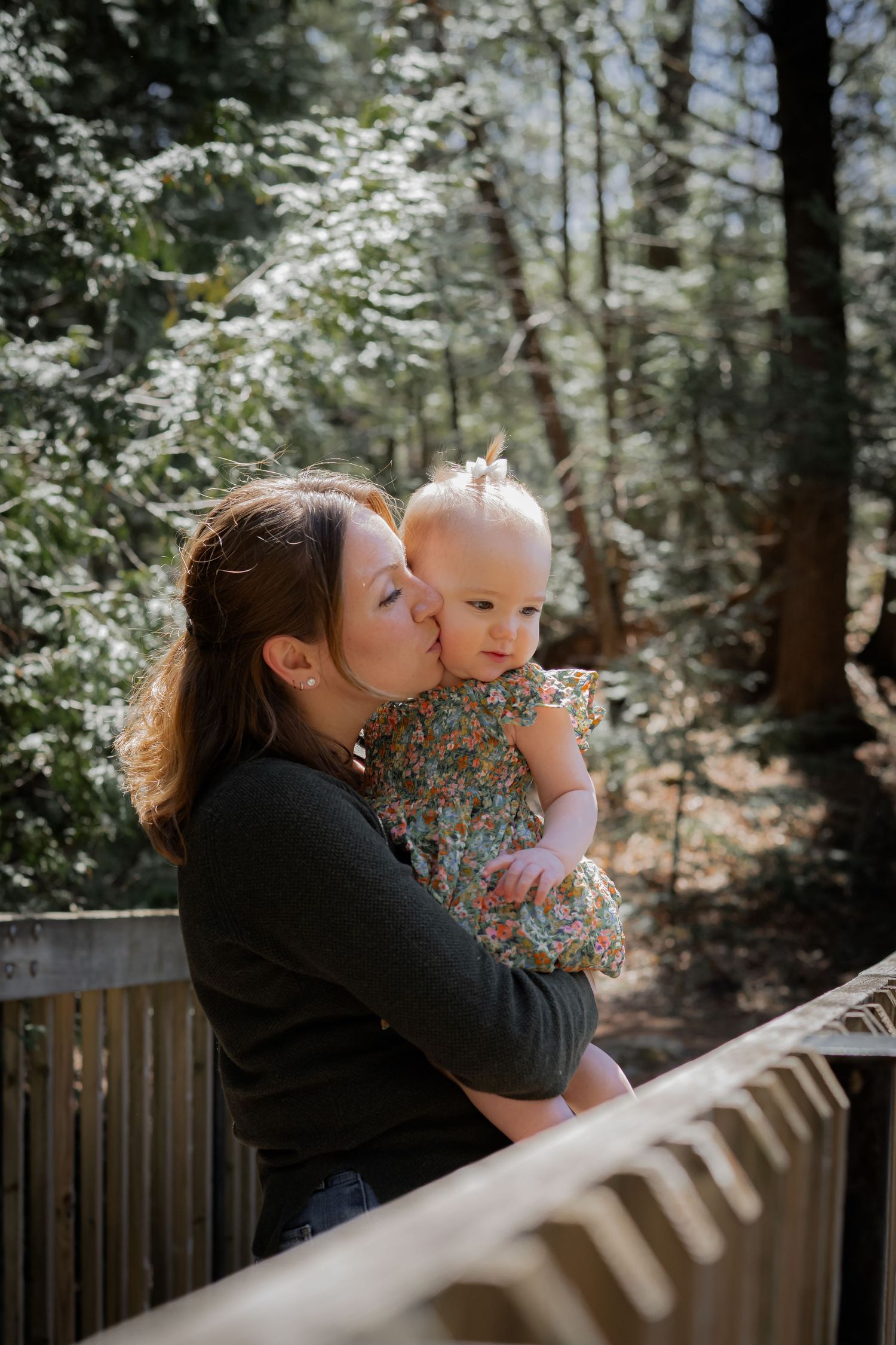 A mother tenderly kisses her baby girl on a wooden deck surrounded by lush green forest trees.