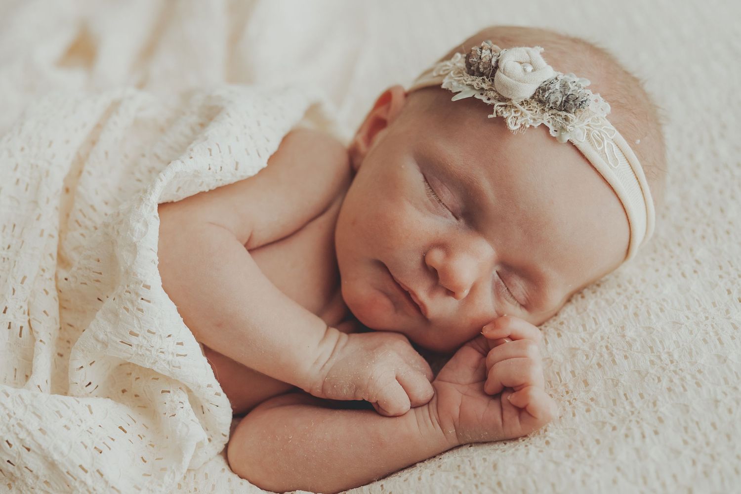 Sleeping newborn in white wrap with delicate lace headband on textured background.