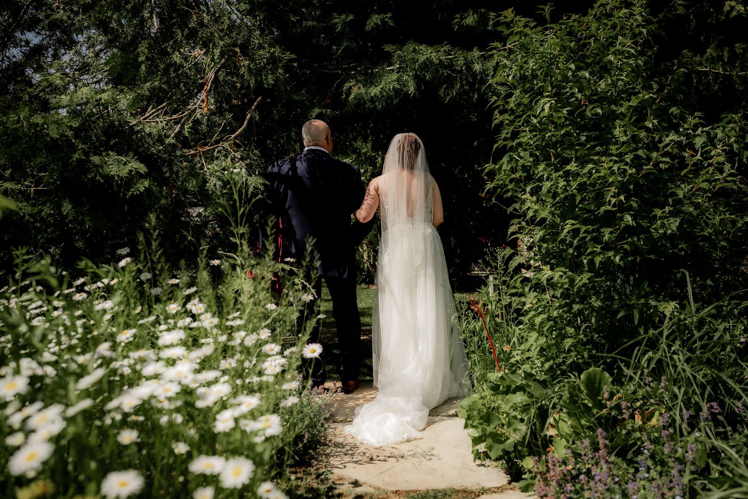 A bride in a flowing white gown walks along a sunlit garden path surrounded by lush greenery and blooming flowers.