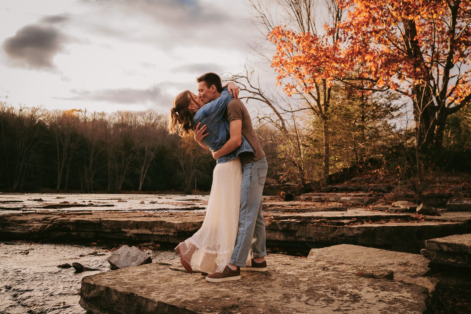 A romantic couple embraces on a rocky outcrop surrounded by autumn trees during a sunset photoshoot.