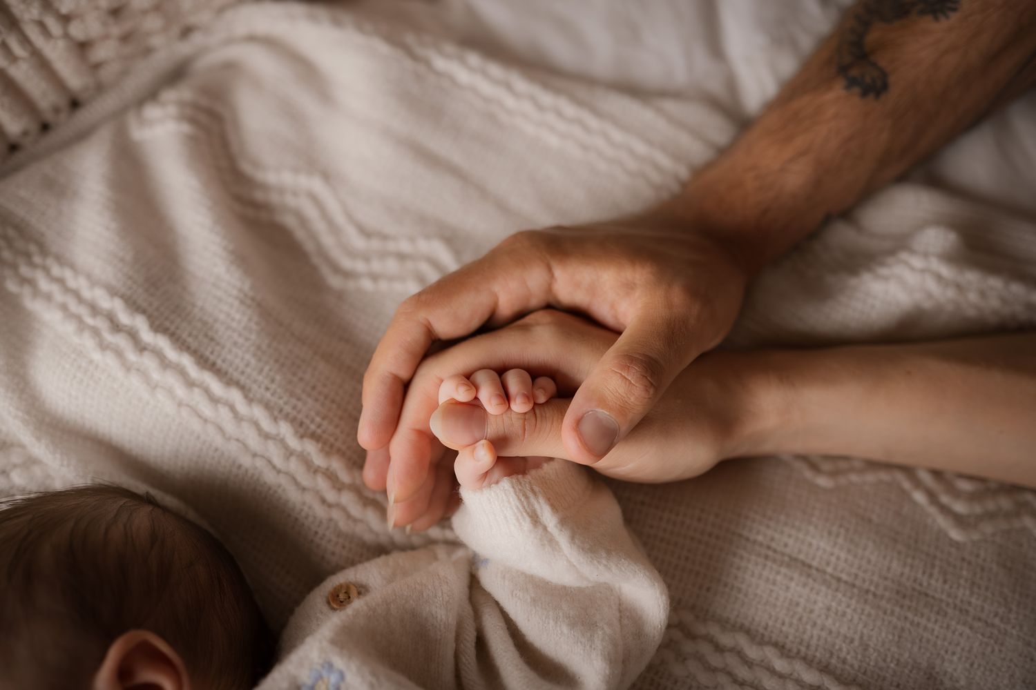 Two adult hands gently cradling a newborn baby's tiny feet on a soft white knit blanket.
