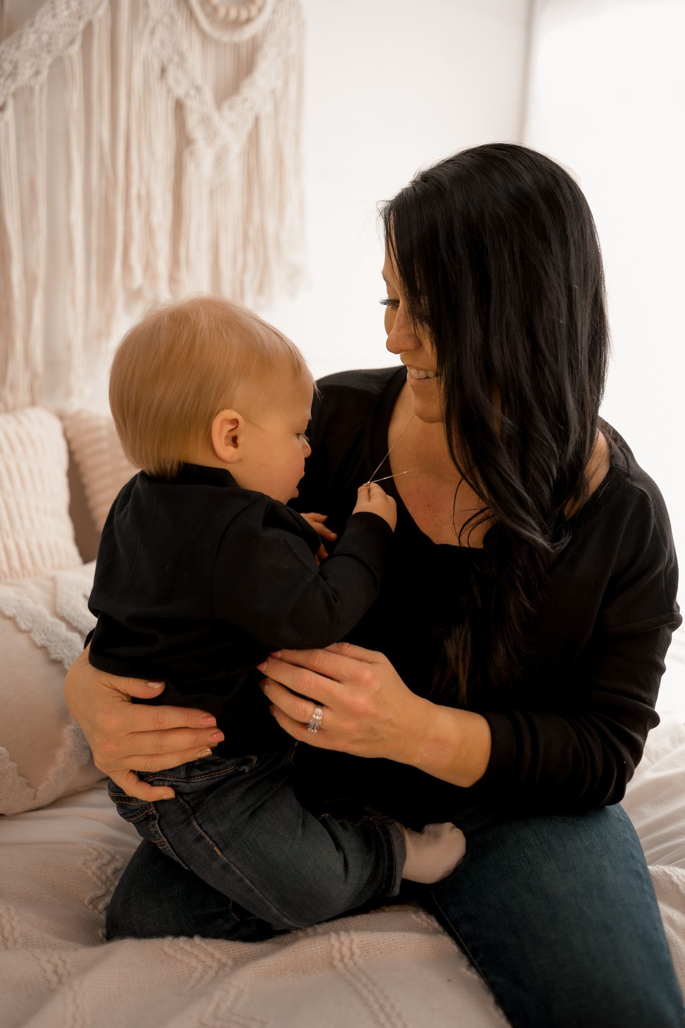 A dark-haired mother in black top holds her baby on a white bed, sharing a tender moment together.