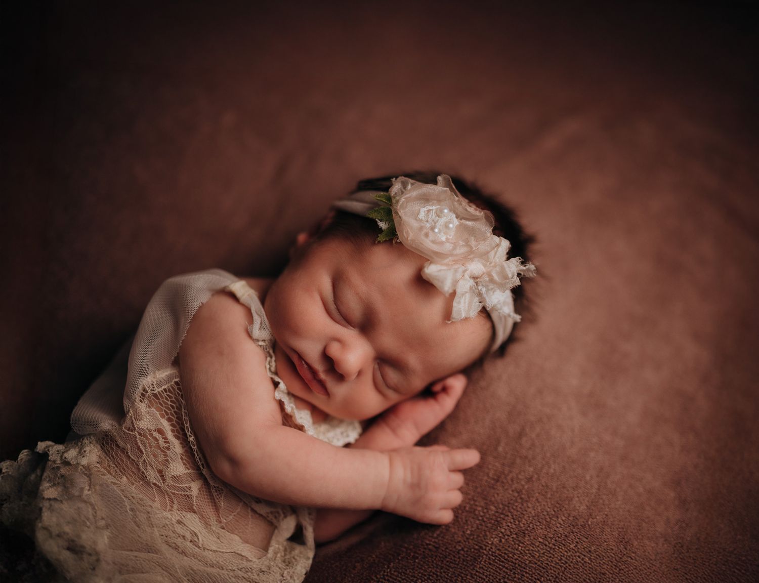 Sleeping newborn baby girl in lace romper and floral headband on brown backdrop.