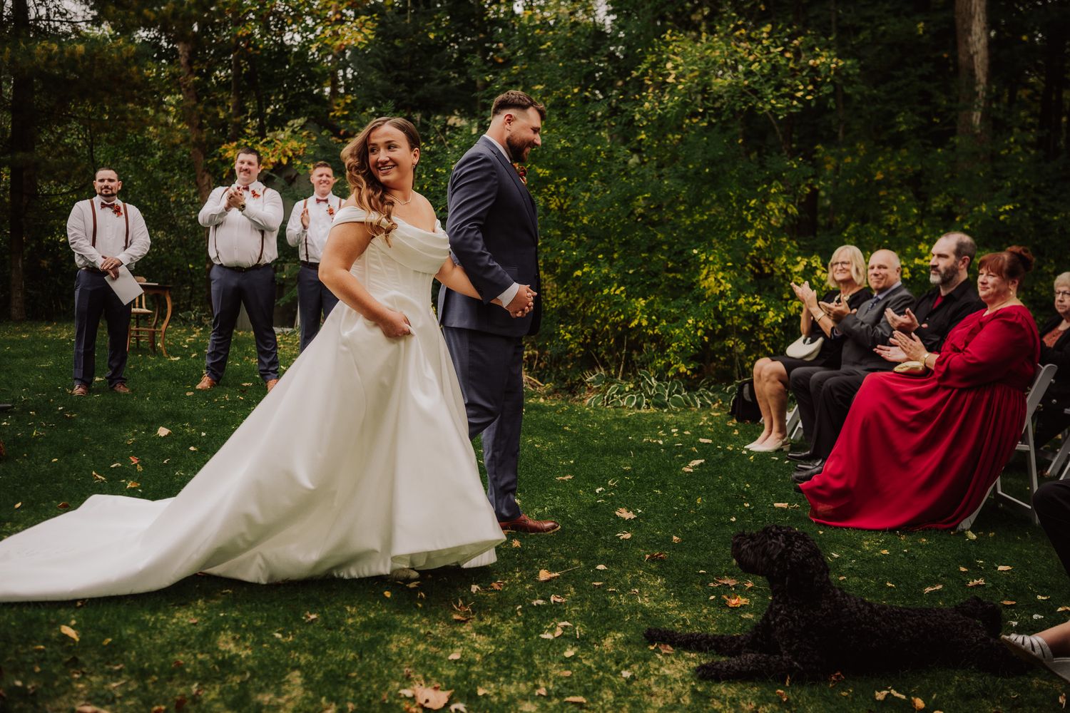 A wedding ceremony takes place outdoors on a lush green lawn with guests seated in chairs during autumn.